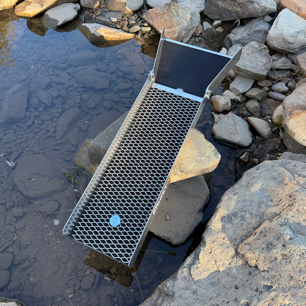 Top side of a 8" sluice , showing the expanded mesh and rubber matting placed outdoors over shallow water between rocks and stones.