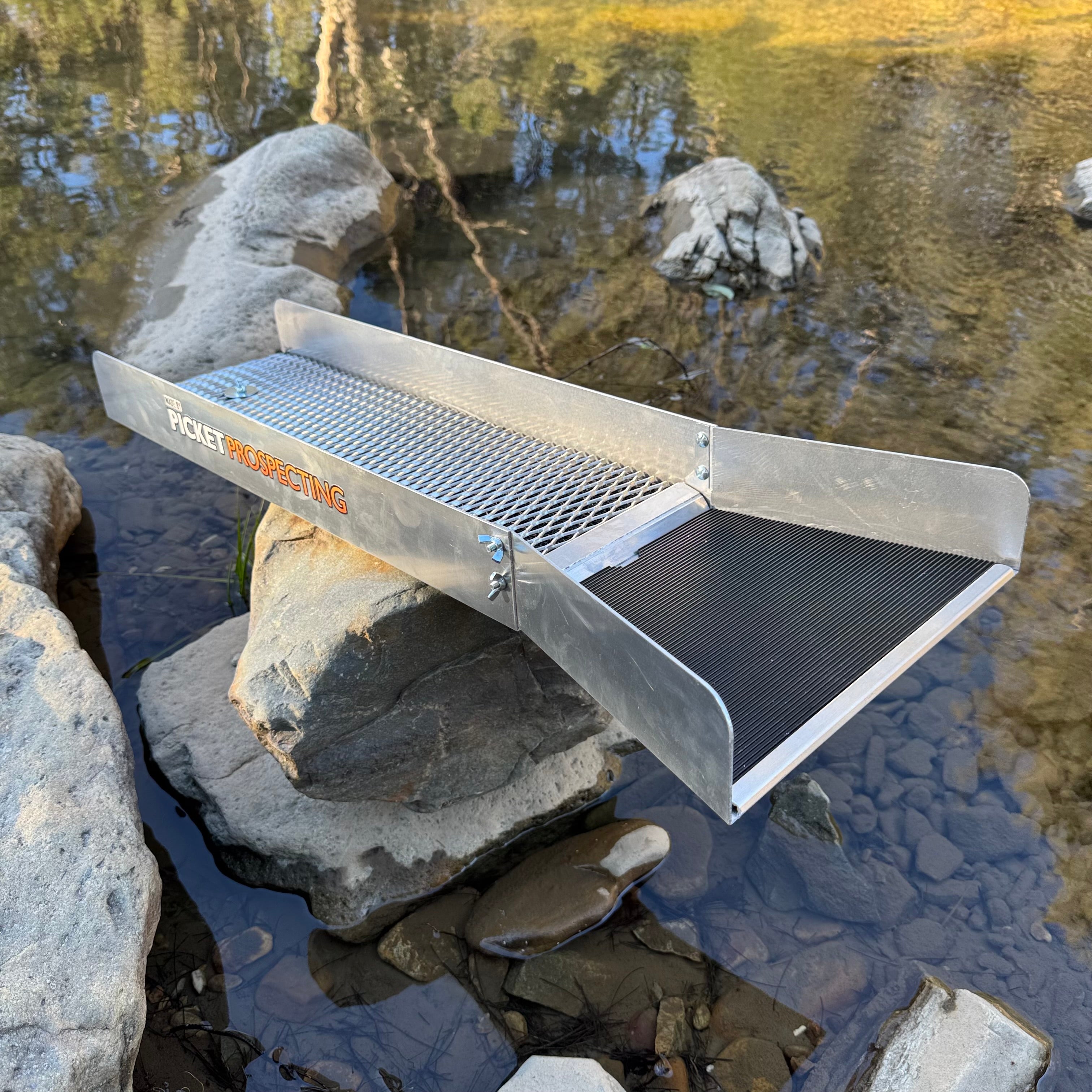 Aluminium sluice with a picket prospecting logo on the side, displaying v matting on the top in a creek setting.