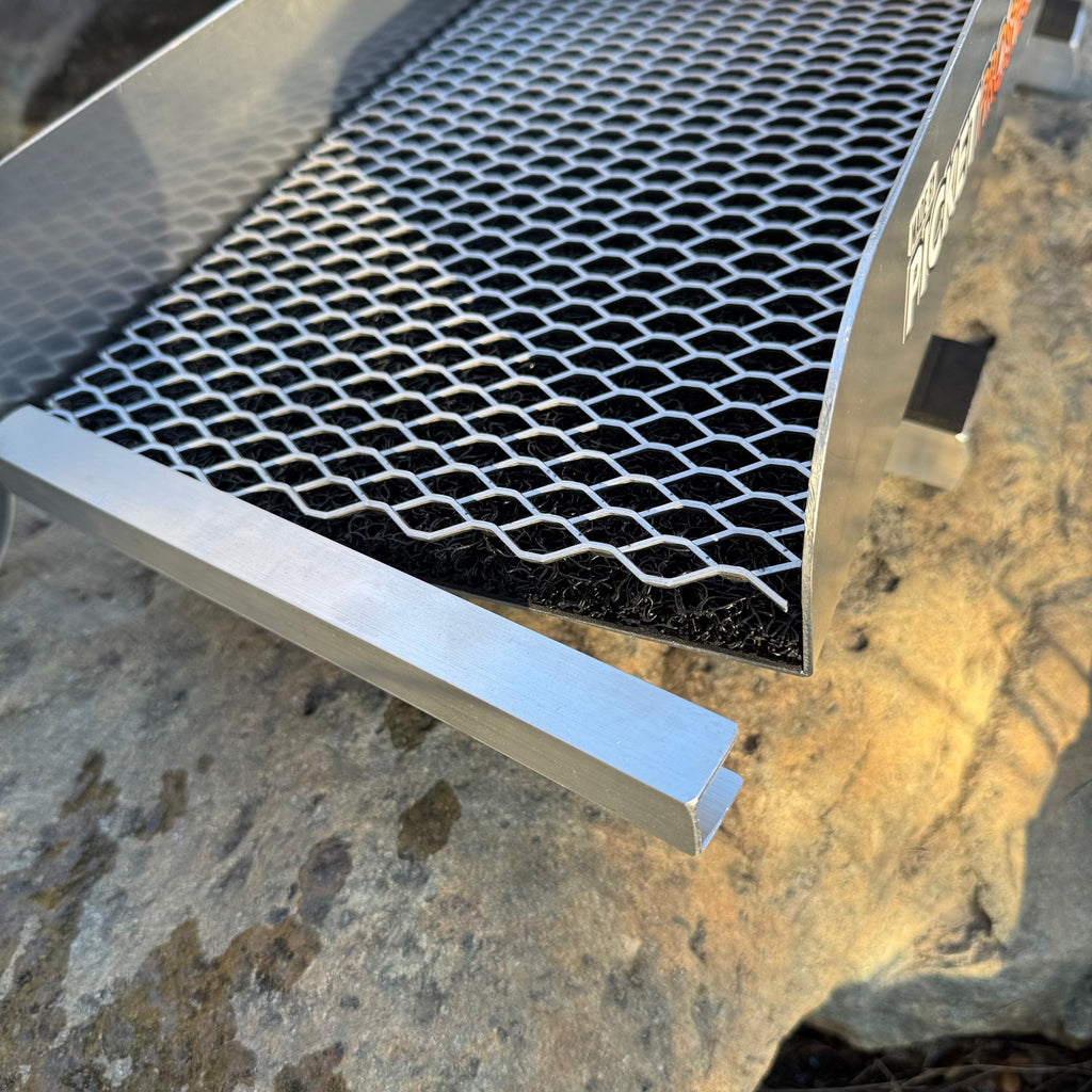 Close-up of a metal grill grate with a black mesh pattern and silver aluminum frame, set outdoors on a stone surface.
