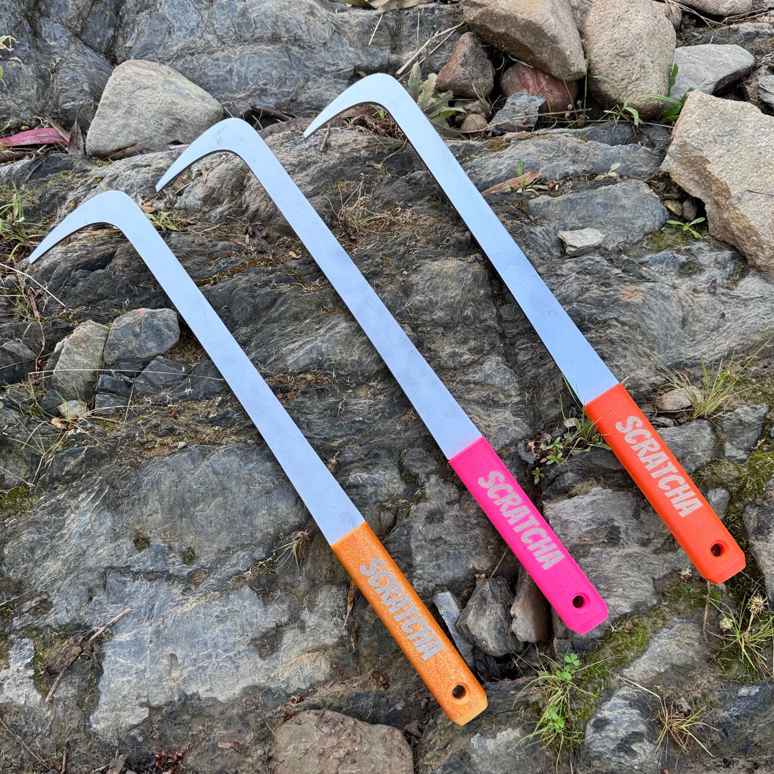 Three long metal scrapers with curved silver blades and colored handles, placed on a rocky outdoor ground. The handles are orange, pink, and red, each printed with the brand name 'SCRAPERS'.