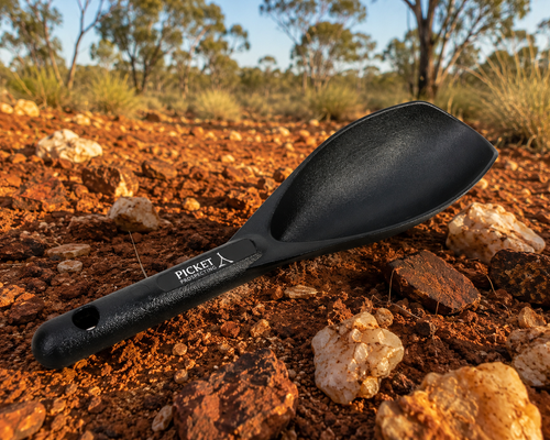 A black Picket brand scoop or shovel with a textured handle, resting on reddish-brown soil and rocks outdoors.