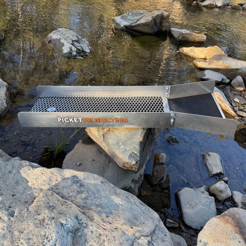 A metal riffle screen or sluice box labeled 'PICKET PROSPECTING' is placed over shallow rocky water in an outdoor stream. The open hop above the riffle and the open hop below are visible, with boulders and flowing water around it.