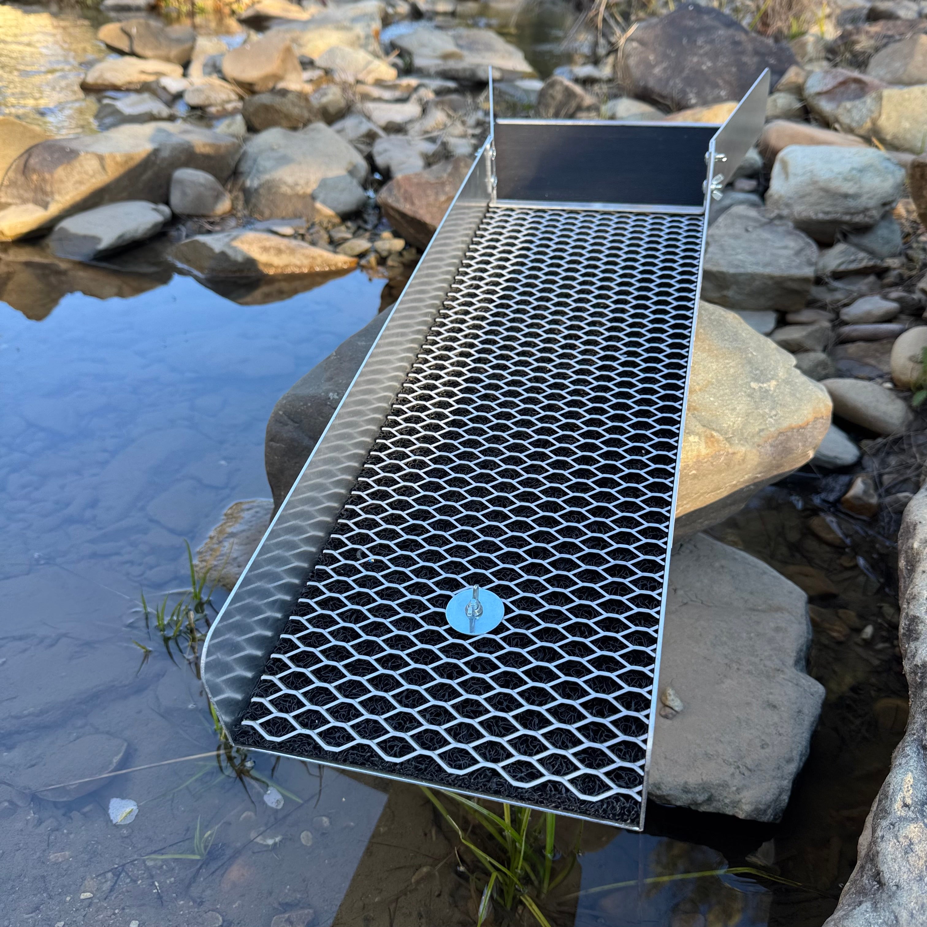 Aluminium sluice with expanded mesh sitting on a large rock in a creek setting.