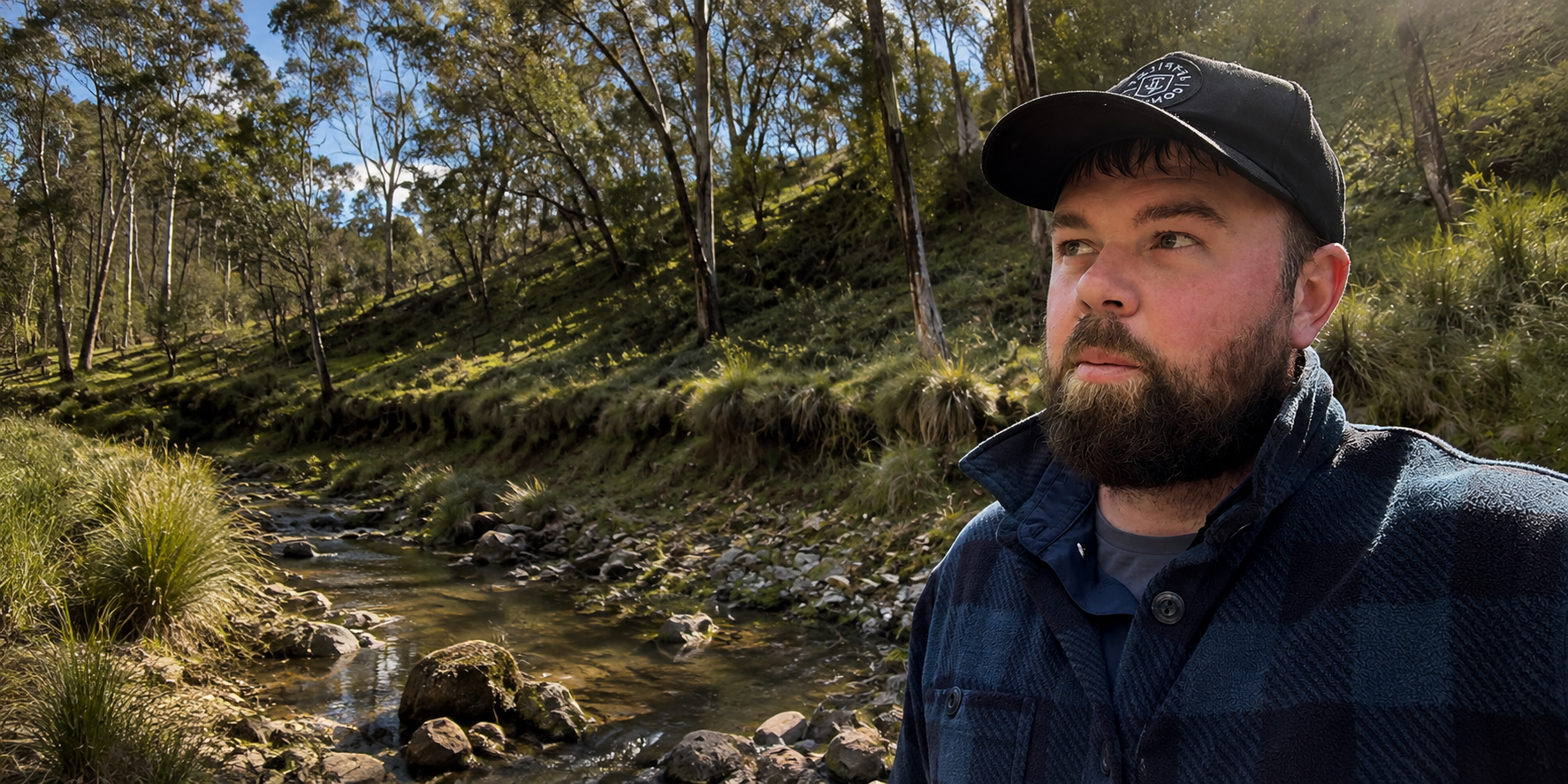 A bearded man wearing a black baseball cap and a dark blue plaid flannel shirt stands outdoors in a natural setting with a small stream, rocks, and a grassy, tree-covered hillside behind him.