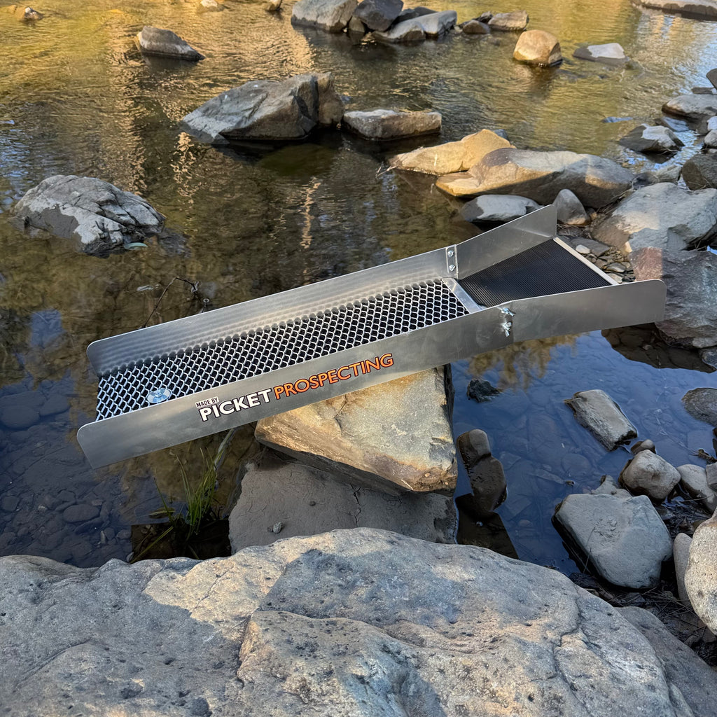 An aluminium sluice with the brand name 'PICKET PROSPECTING' printed on it, positioned over a rocky stream.