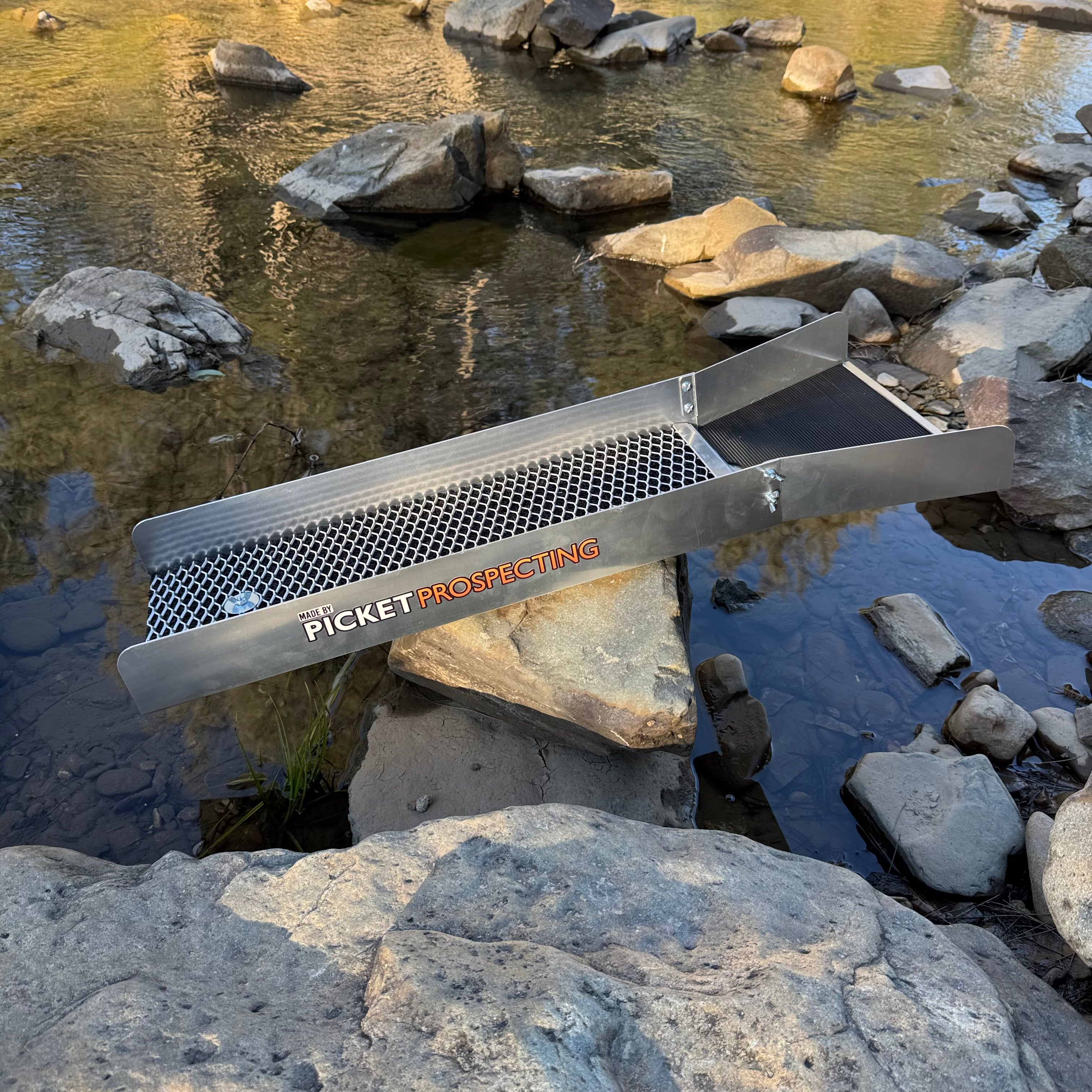An aluminium sluice with the brand name 'PICKET PROSPECTING' printed on it, positioned over a rocky stream.