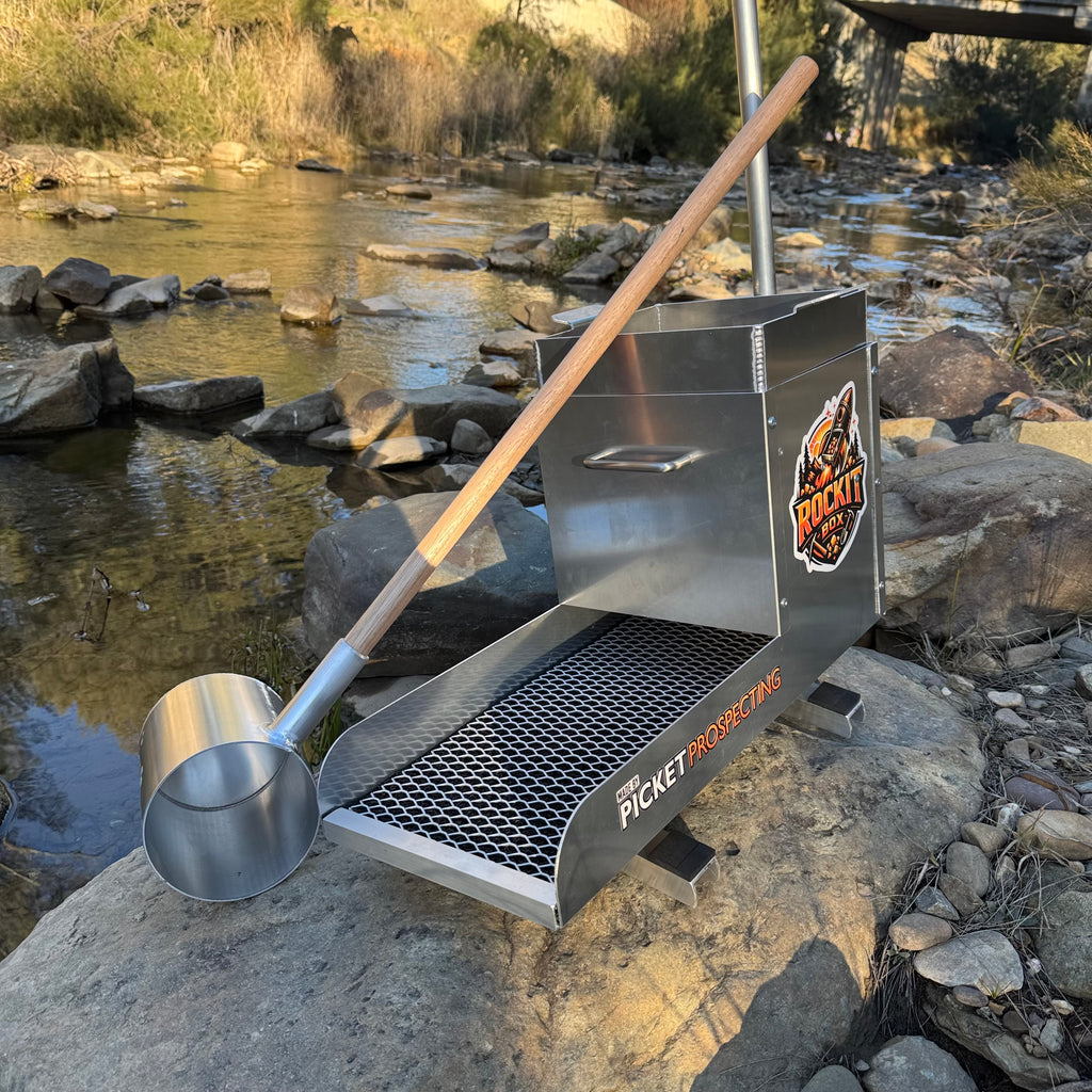 Aluminium rocker box placed on a large rock in a creek setting. With aluminium water scoop with a timber handle. Displaying expanded mesh and a classifying sieve.
