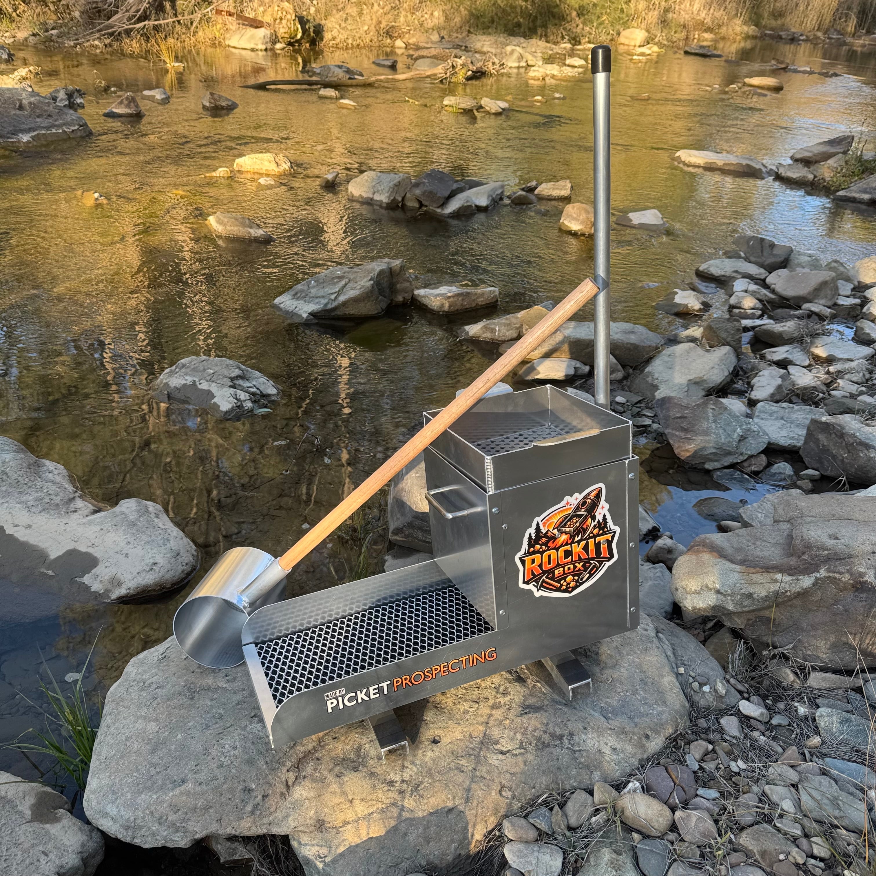 Aluminium rocker box placed on a large rock in a creek setting.Scoop resting on the front of the box. Displaying expanded mesh and a classifying sieve.