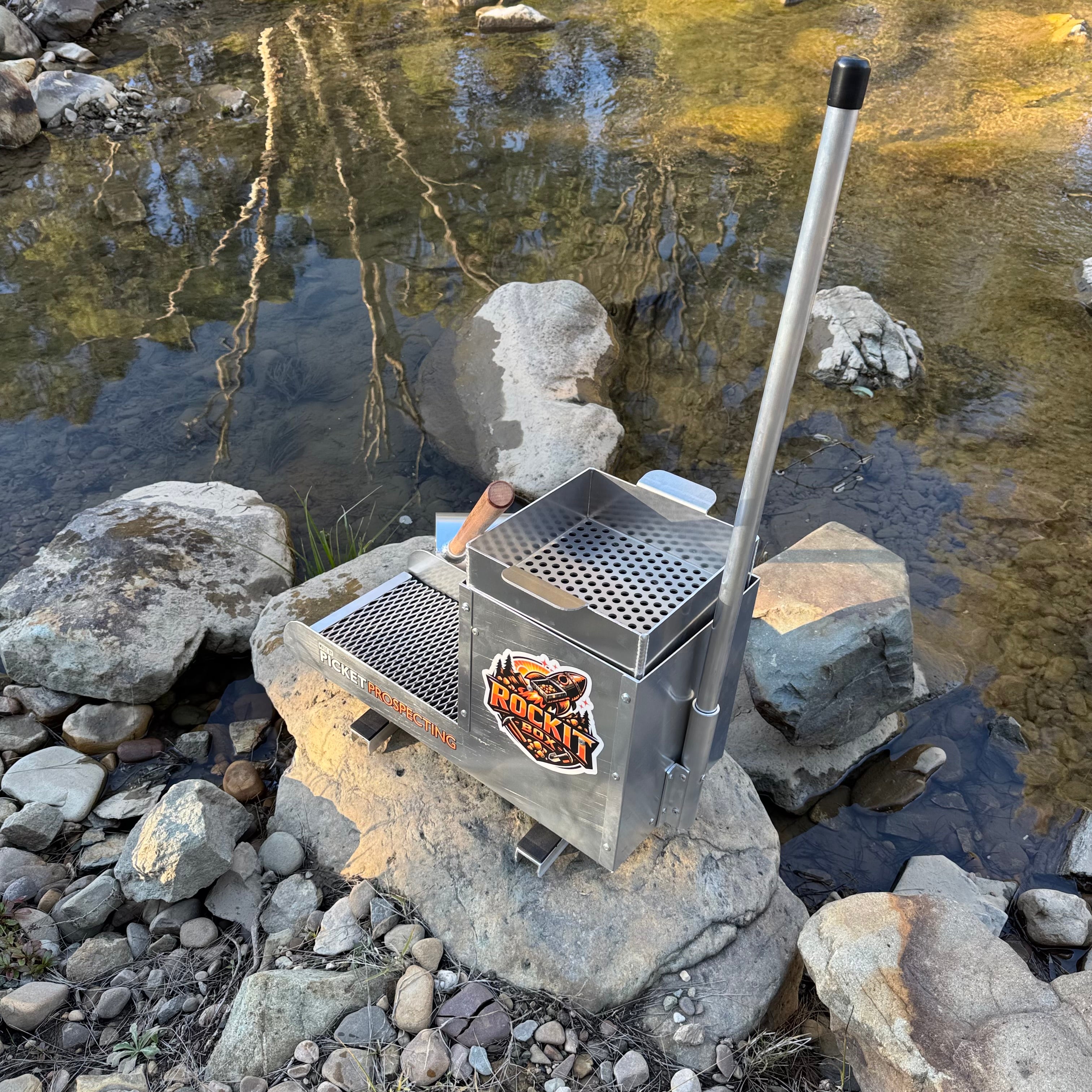 Aluminium rocker box placed on a large rock in a creek setting. Displaying expanded mesh and a classifying sieve.