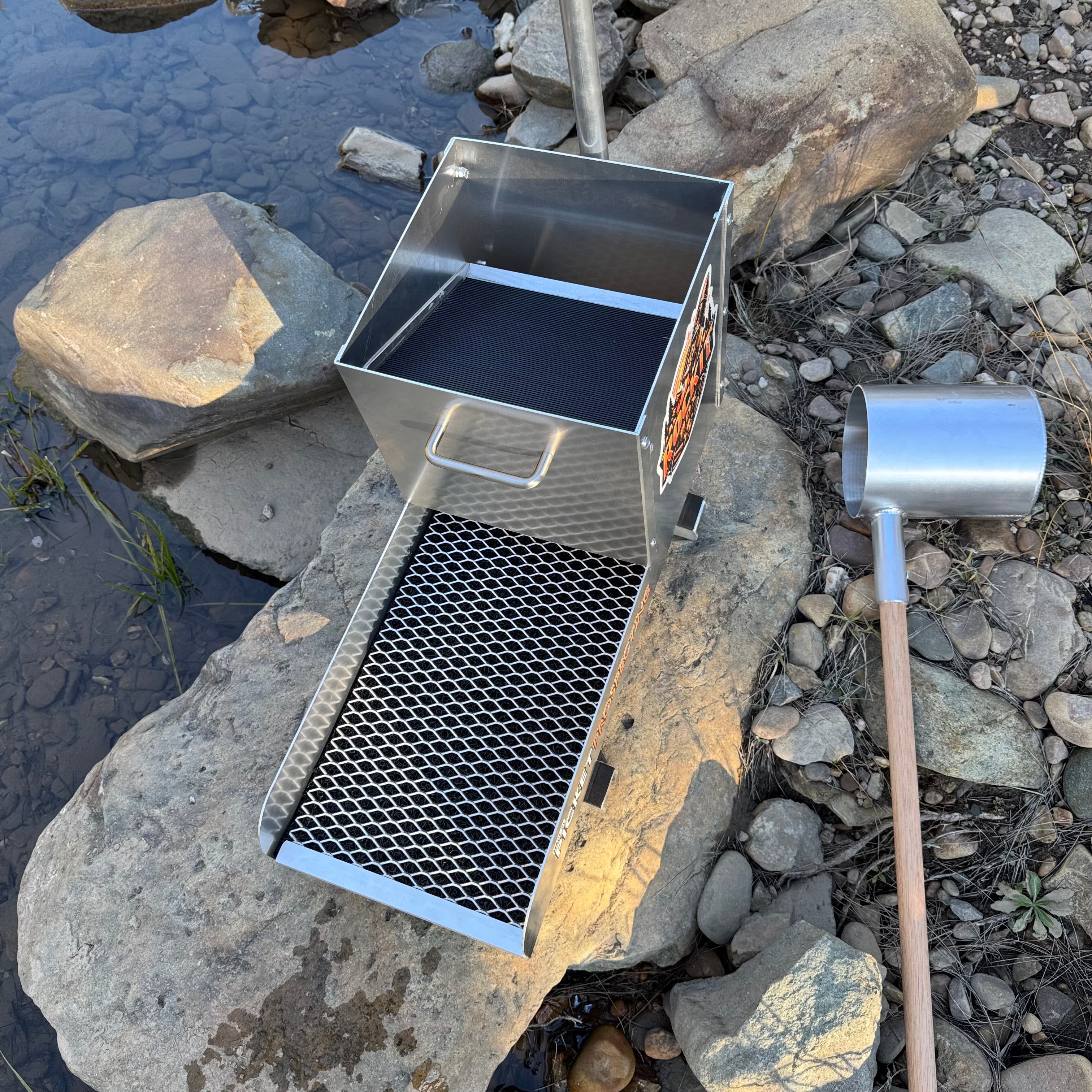 Aluminium rocker box placed on a large rock in a creek setting. Displaying expanded mesh and a classifying sieve.