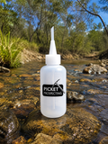 A translucent white plastic bottle with a pointed nozzle cap stands on a wet rock in a shallow stream outdoors. The bottle has a black label reading 'PICKET PROSPECTING' with a pickaxe logo.