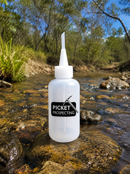 A translucent white plastic bottle with a pointed nozzle cap stands on a wet rock in a shallow stream outdoors. The bottle has a black label reading 'PICKET PROSPECTING' with a pickaxe logo.